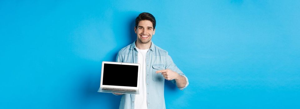 Handsome young man pointing finger at screen of computer, smiling pleased, showing promo in internet or website, standing over blue background.