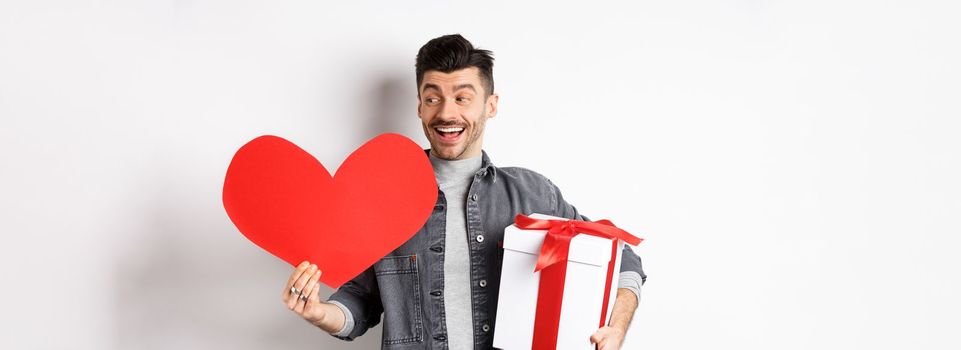 Happy boyfriend making romantic gifts on Valentines day date, holding box with present and big red heart card, smiling cheerful, white background.