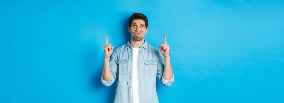 Portrait of doubtful adult man looking disappointed, pointing fingers up at something with uncertain face, standing over blue background.