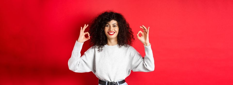 Confident pretty girl with curly hairstyle, showing okay gestures and smiling, approve and agree with you, praising excellent choice, standing satisfied on red background.