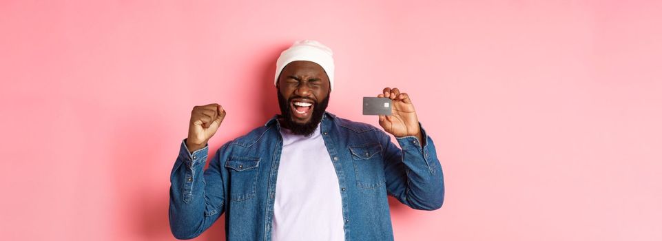 Shopping concept. Happy Black man rejoicing, scream of joy and showing credit card, standing over pink background.