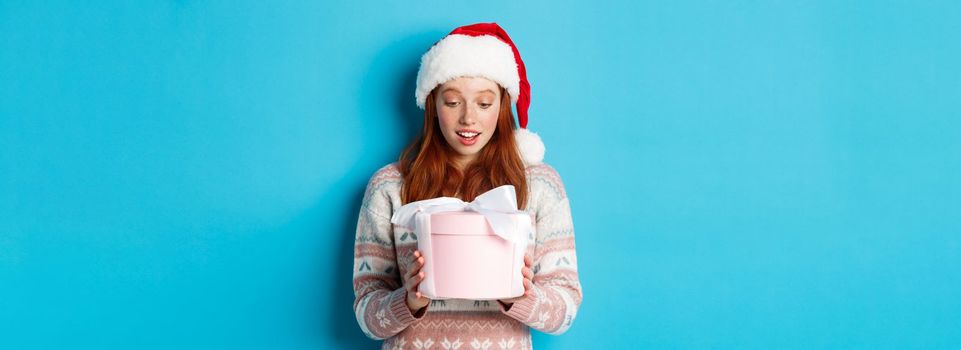 Winter and Christmas Eve concept. Touched and flattered redhead girl looking at box with xmas gift, smiling amazed, standing in santa hat against blue background.