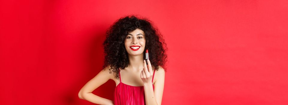 Fashionable woman with curly hair, showing red lipstick and smiling, recommend cosmetic, standing on white background.