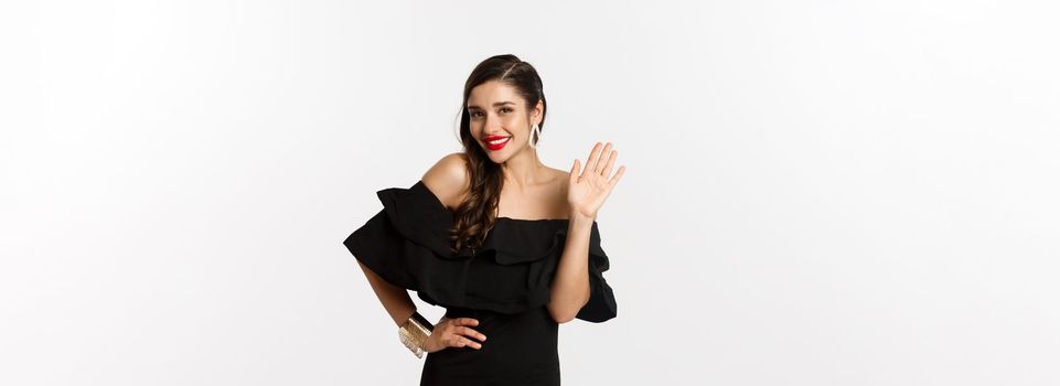 Gorgeous young woman in black dress, earrings and jewelry, waving hand and smiling friendly, saying hello, standing over white background.