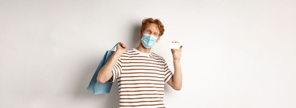 Concept of covid-19 and lifestyle. Cheerful young man with red hair, wear medical mask, showing shopping bag from store and plastic credit card.