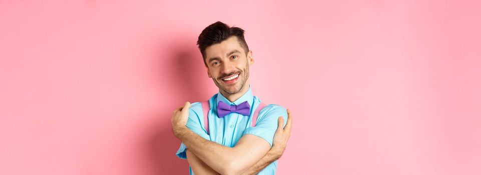 Cheerful young man smiling and hugging himself, wrap hands around own body and looking happy, standing over pink background.