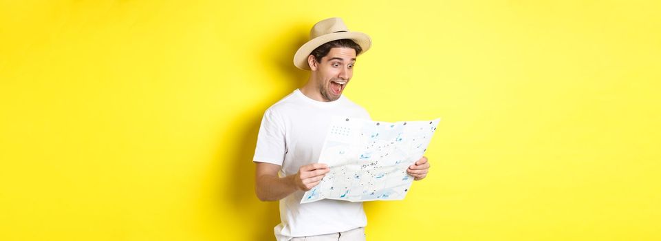 Travelling, vacation and tourism concept. Smiling happy tourist looking at map with sightseeings, standing against yellow background.