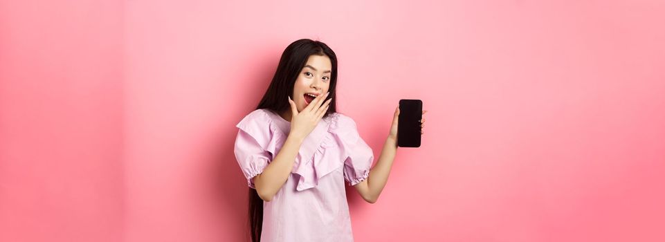 Cheerful asian teen girl showing empty phone screen, laughing and covering mouth with hand, standing in dress against pink background.