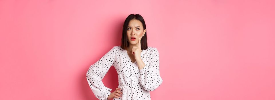 Thoughtful asian woman making assumption, looking up and thinking, deciding something, standing in dress over pink background.