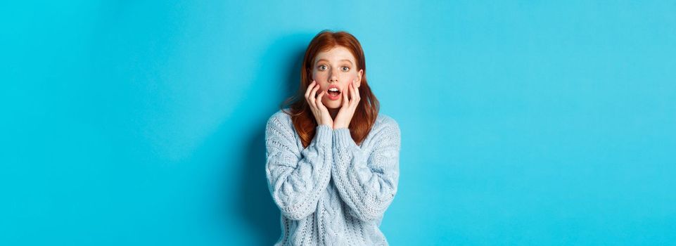 Startled redhead girl staring at something amazing, gasping in awe, standing in sweater against blue background. Copy space