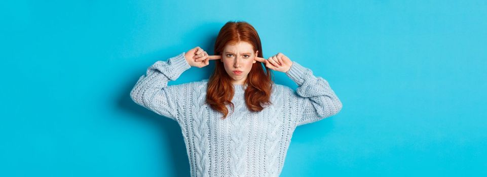 Teenage redhead girl unwilling to listen, shut ears and frowning angry, staring at camera offended, sulking against blue background.