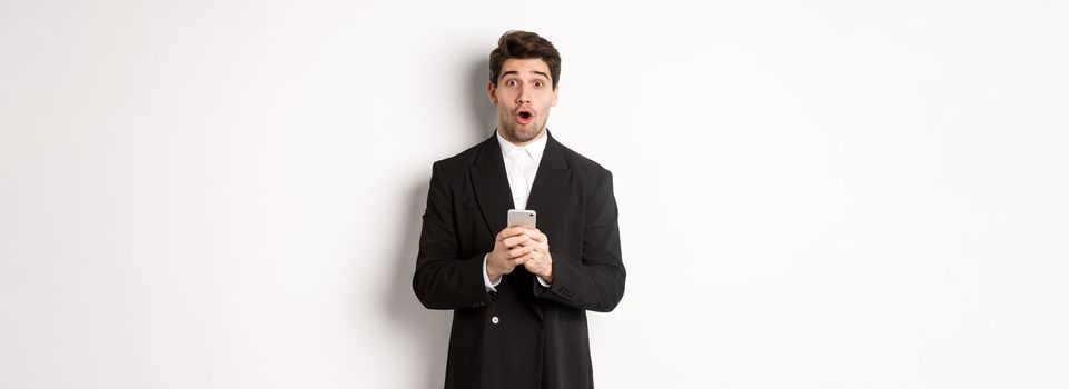 Amazed handsome guy in black suit reacting to cool promo offer, holding mobile phone, standing against white background.