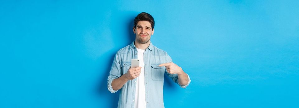 Concept of online shopping, applications and technology. Skeptical and displeased guy pointing finger at smartphone and grimacing disappointed, standing over blue background.