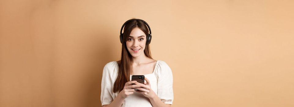 Pretty girl in headphones smiling at camera, listening music and using mobile phone app, beige background.