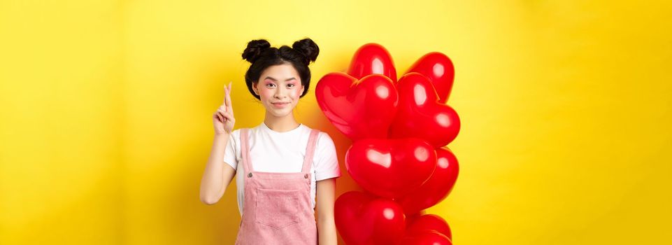 Valentines day concept. Stylish teenage asian girl with romantic make-up look, holding fingers crossed for good luck, making wish and smiling, standing on yellow background.