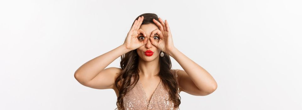Concept of New Year celebration and winter holidays. Close-up of beautiful brunette woman in dress, red lips, making hand binoculars and staring at camera, white background.