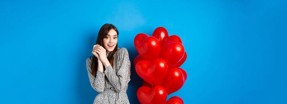 Valentines day. Lovely girlfriend ind ress smiling happy at camera, celebrating lovers holiday, standing near heart balloons, blue background.