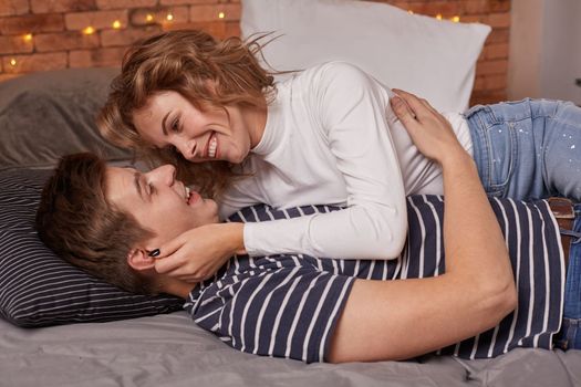 Caucasian young beautiful couple lie on bed and hugging each other. They look each other in the eyes and they are happy