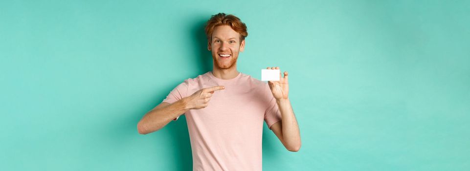 Attractive adult man with beard and red hair pointing finger at plastic credit card, smiling pleased at camera, standing over turquoise background.
