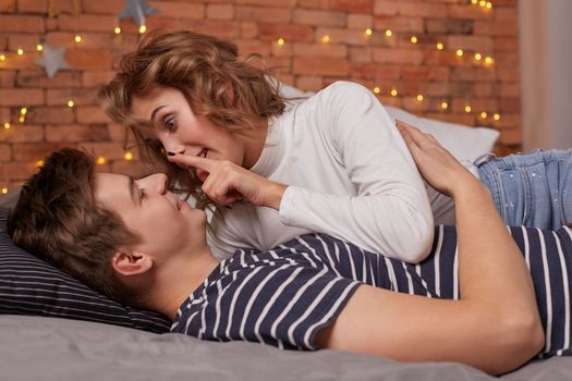 Caucasian young beautiful couple lie on bed and hugging each other. They look each other in the eyes and they are happy