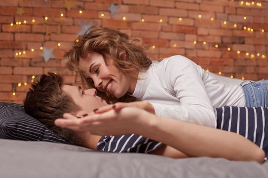 Caucasian young beautiful couple lie on bed and hugging each other. They look each other in the eyes and they are happy