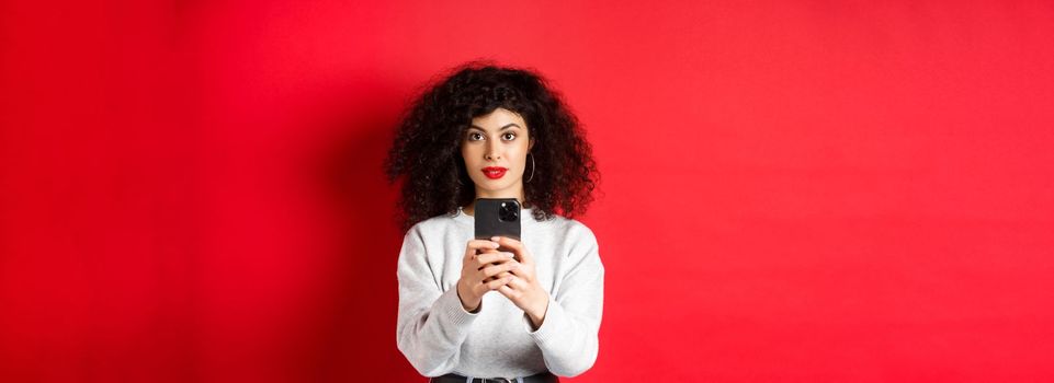 Young woman with curly hair, recording video on smartphone, taking photo on mobile phone and looking at camera, standing on red background.