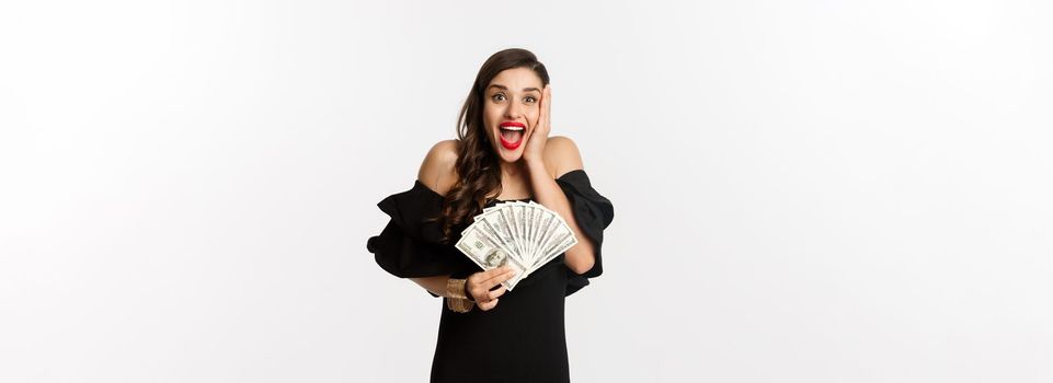 Fashion and shopping concept. Attractive woman rejoicing of money prize, holding dollars and shouting from excitement, standing in black dress over white background.