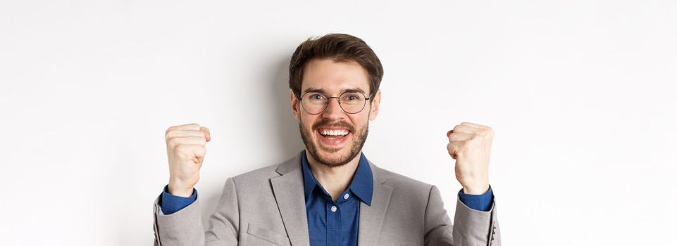 Close-up of cheerful male ceo manager in glasses and suit, saying yes and celebrating win, making fist pump, standing on white background.