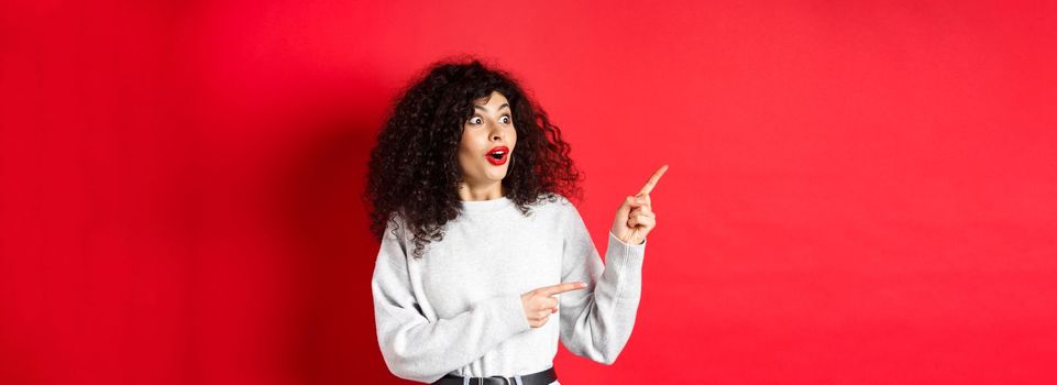 Surprised attractive girl with curly hair, pointing and looking aside at empty space, showing logo or advertisement, standing on red background.