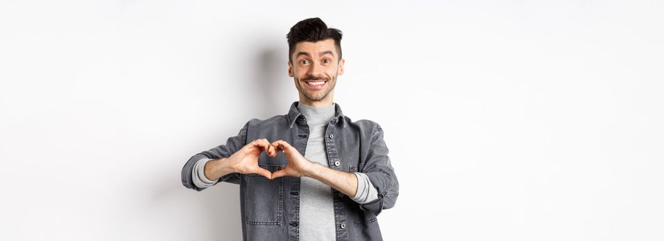 Smiling boyfriend showing heart with love, looking romantic and happy at lover, standing on white background. Valentines day and relationship concept.