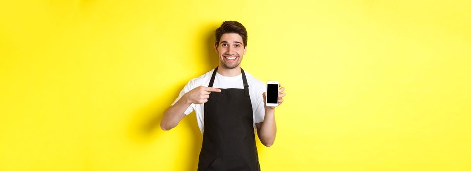 Handsome barista in black apron pointing finger at mobile screen, showing app and smiling, standing over yellow background.