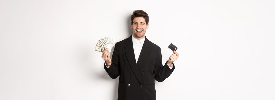 Image of confident businessman in black suit, smiling pleased and winking, holding money and credit card, standing against white background.