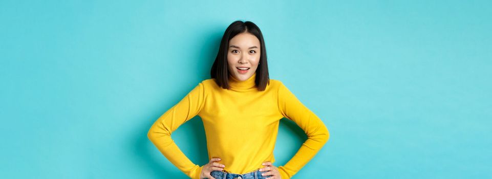 Image of impressed korean girl listening interesting story, looking at camera intrigued, standing over blue background.