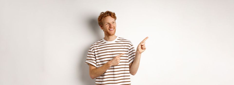 Cheerful caucasian guy with red hair and beard, looking and pointing left at logo banner, standing over white background.