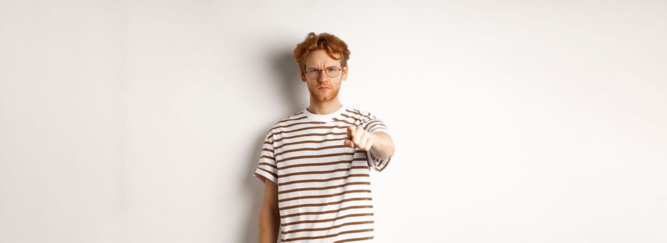 Angry and serious guy with red hair accusing you, frowning and pointing finger at camera, blame someone, standing over white background.