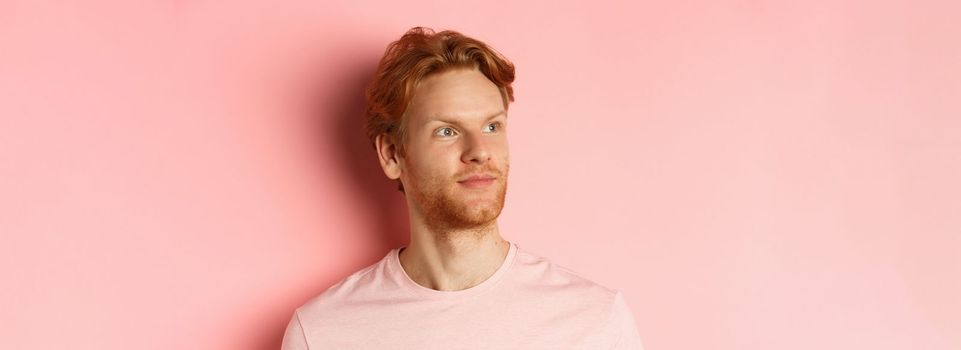 Handsome european male model with red hair and beard, turn head and looking pleased at copy space on left side, standing over pink background.