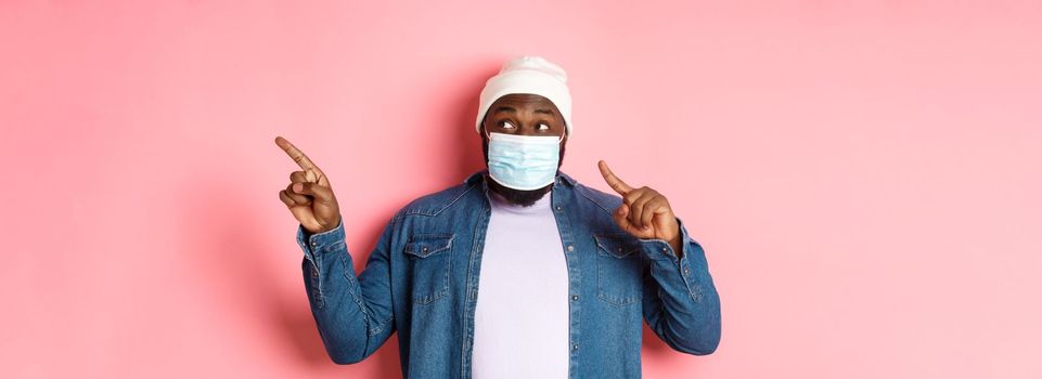 Covid-19, lifestyle and lockdown concept. Amused Black man in medical mask, looking and pointing right at copy space, standing over pink background.