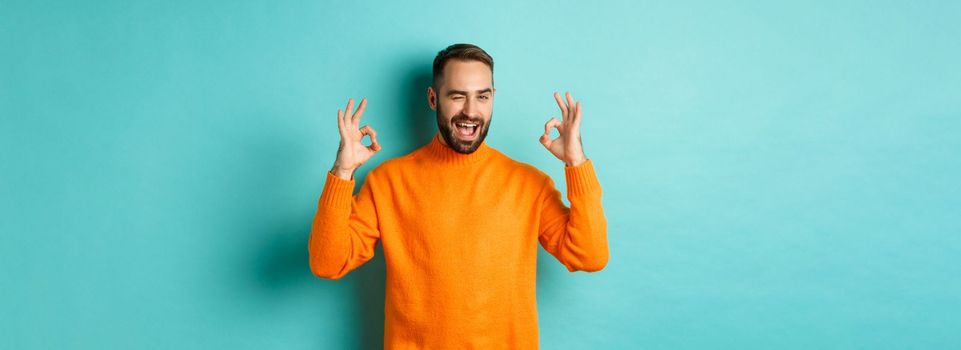 Confident man assuring all ok, showing okay signs and winking at you, recommending product, standing in orange sweater at studio background.