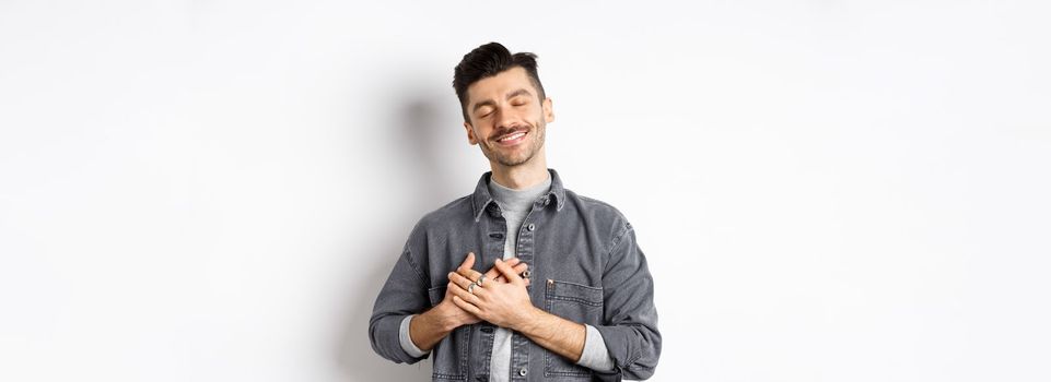 Romantic happy man holding hands on heart and smiling with closed eyes, dreaming of something, feeling nostalgic from sweet memories, standing on white background.
