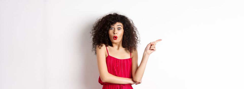 Surprised elegant woman in red dress, saying wow and pointing finger left, showing interesting promo, standing over white background.