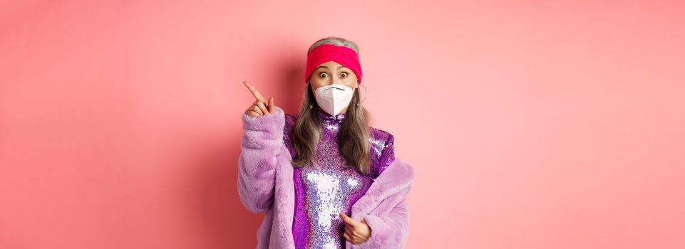 Coronavirus and shopping concept. Amazed asian senior woman in face mask staring at camera, pointing upper right corner promo, standing over pink background.