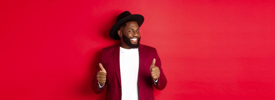 Cheerful Black man having fun on party, showing thumbs up in approval, smiling and liking something, standing against red background.