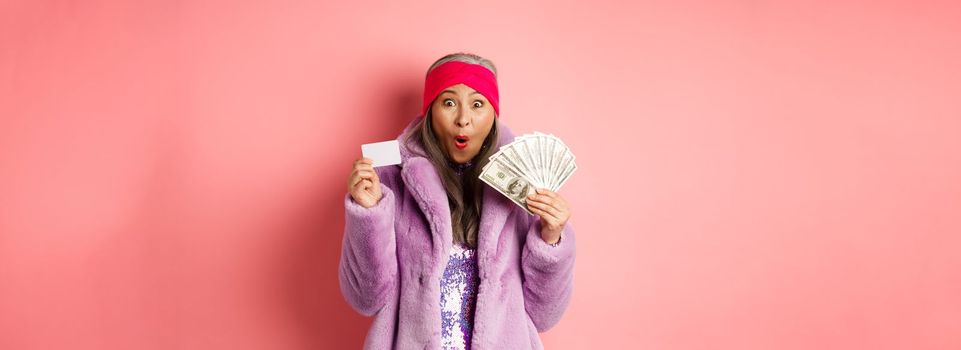 Shopping and fashion concept. Happy senior asian woman holding money in dollars and plastic card, looking amazed and ecstatic, pink background.