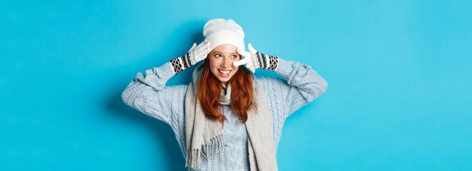 Winter and holidays concept. Cute redhead teen girl in beania, gloves and sweater showing peace sign, looking left at copy space on blue background.