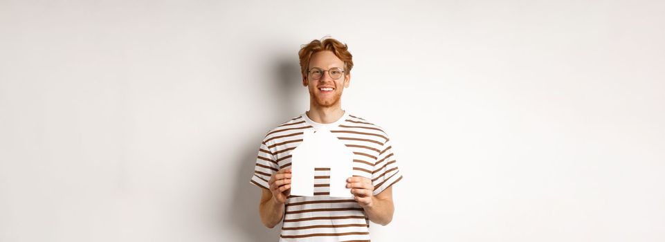 Handsome young man with beard and red hair, wearing glasses and striped t-shirt, showing paper house cutout and smiling, concept of real estate and buying property.