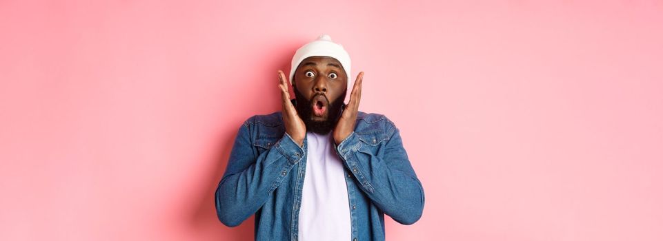 Shocked and impressed Black man staring at camera with complete disbelief, saying wow, standing in beanie and hipster shirt over pink background.