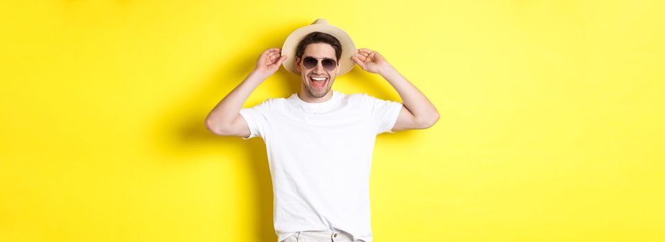 Happy man on vacation, wearing straw hat and sunglasses, smiling while standing against yellow background.