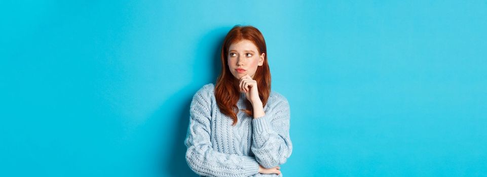 Thoughtful and upset redhead girl looking right, pondering solution, standing in sweater against blue background.