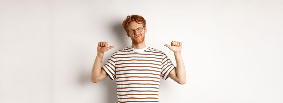 Confident young nerdy guy in glasses with red hair, pointing at himself and looking like professional, white background.