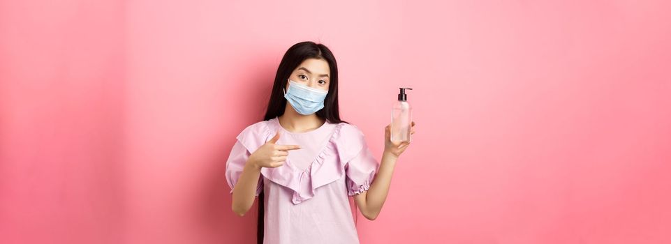 Healthy people and covid-19 pandemic concept. Stylish japanese girl in medical mask showing bottle of hand sanitizer, pointing at antiseptic, standing against pink background.